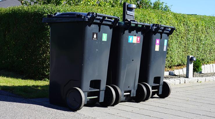Tre affaldsbeholdere med piktogrammer står i skel i indkørsel med grøn hæk i baggrunden